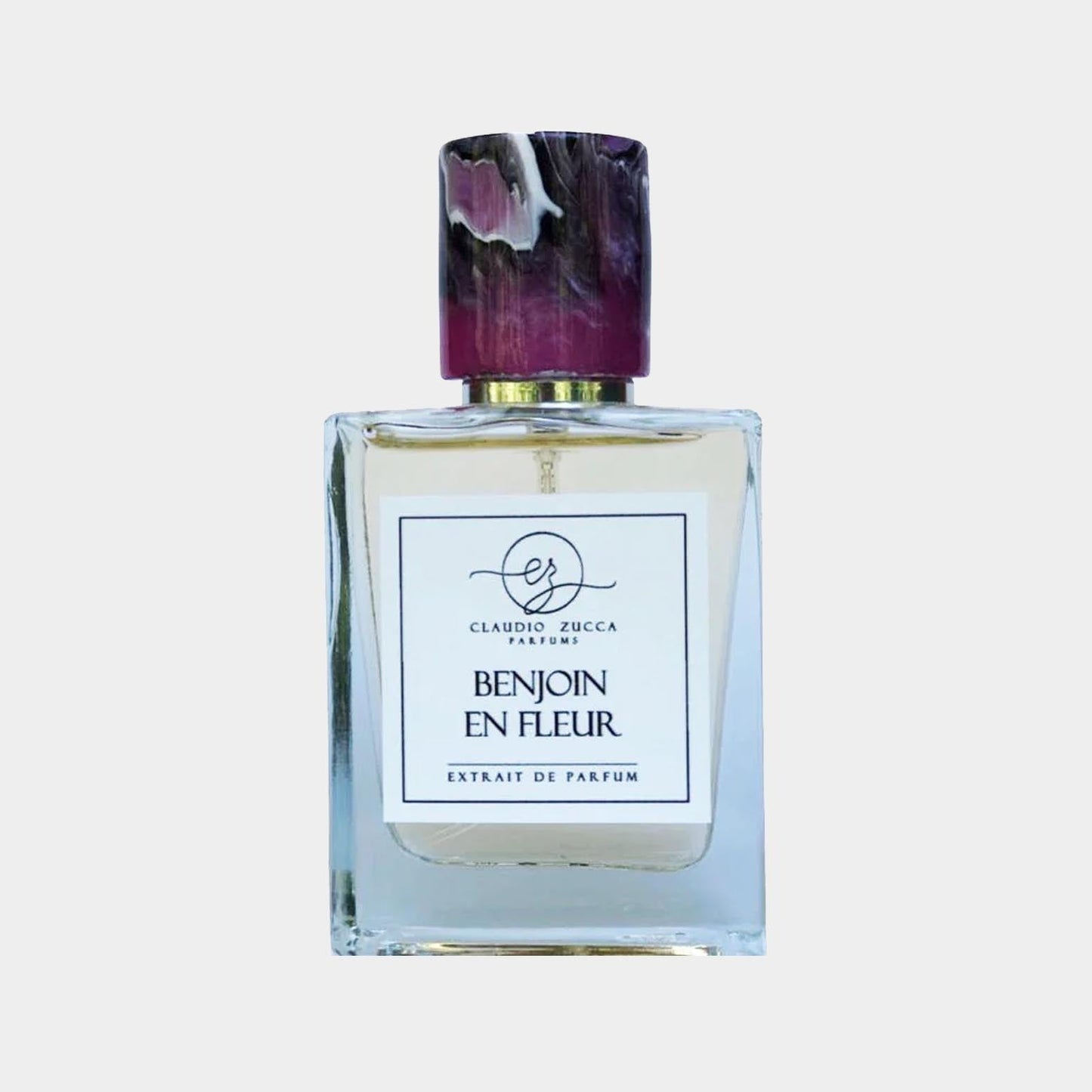 A full bottle of Claudio Zucca Benjoin en Fleur, featuring a clear glass design and a striking purple-and-gold cap, labeled as an extrait de parfum. The elegant bottle showcases the fragrance's refined aesthetic.