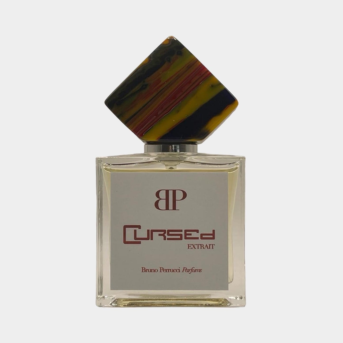 A stylish bottle of Bruno Perrucci Cursed Sample perfume, featuring a clear glass design and a distinctive multi-colored cap. The label reads Cursed Extrait in elegant lettering.
