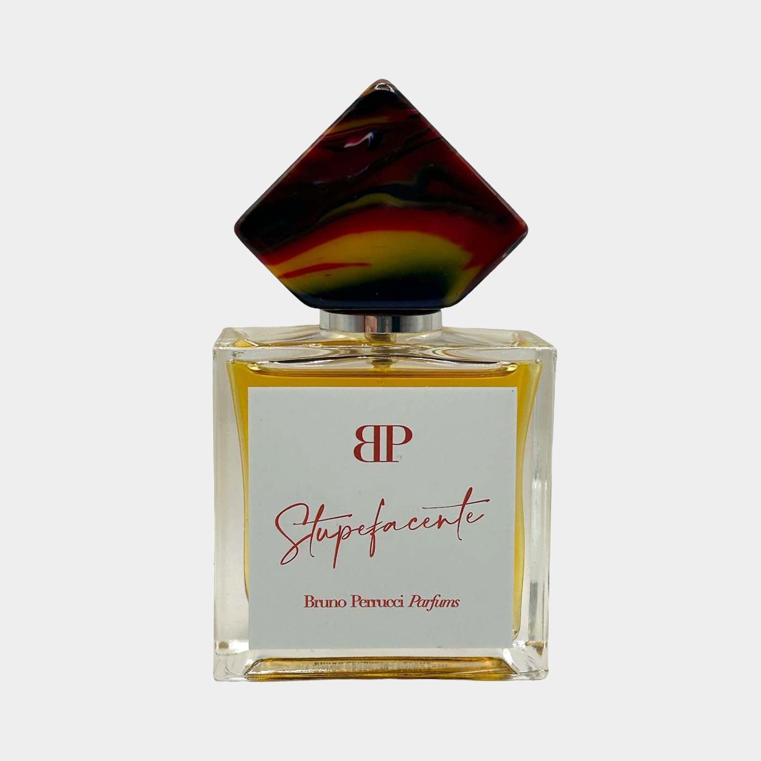 A full bottle of Bruno Perrucci Stupefacente perfume, featuring a square glass base and a uniquely shaped cap with vibrant colors. The elegant label displays the brand name and fragrance title.