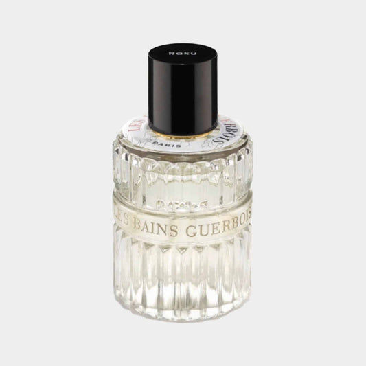 A clear glass bottle of Les Bains Guerbois Raku EDP, featuring a textured design and a black cap, sitting elegantly on a light background. The label displays the brand name and fragrance details.