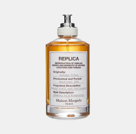 A bottle of Maison Martin Margiela Replica Autumn Vibes Sample, featuring a warm amber liquid and a label describing its woody, floral, and red leaf fragrance, evoking the essence of autumn in Montreal.
