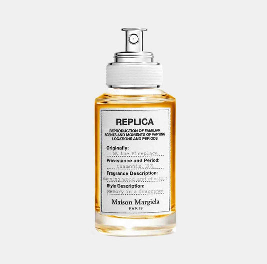 A bottle of Maison Martin Margiela Replica By the Fireplace fragrance sample, featuring a clear glass design with a silver spray nozzle and a label detailing its scent profile. The fragrance evokes warmth with notes of burning wood and chestnut.