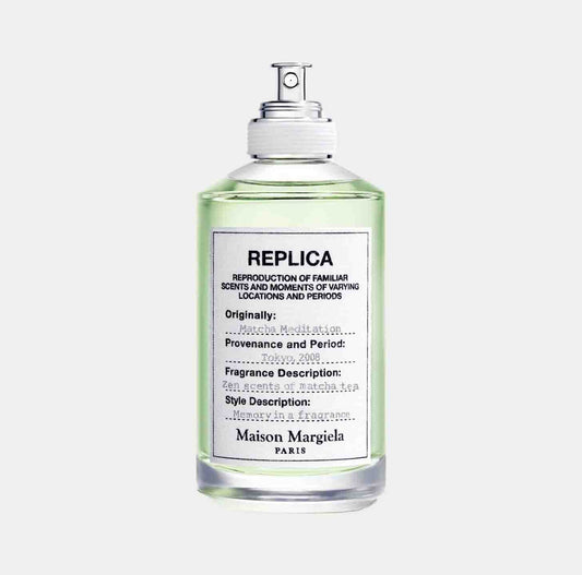 A bottle of Maison Martin Margiela Replica Matcha Meditation Sample, featuring a light green liquid and a label that describes its fragrance and origins. The scent captures zen moments with notes of matcha tea.
