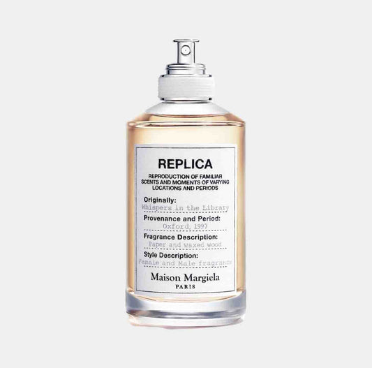 A clear glass bottle of Maison Martin Margiela Replica Whispers in the Library sample fragrance, featuring a minimalist label with fragrance notes and details. The liquid inside is a warm amber color, evoking a cozy atmosphere.