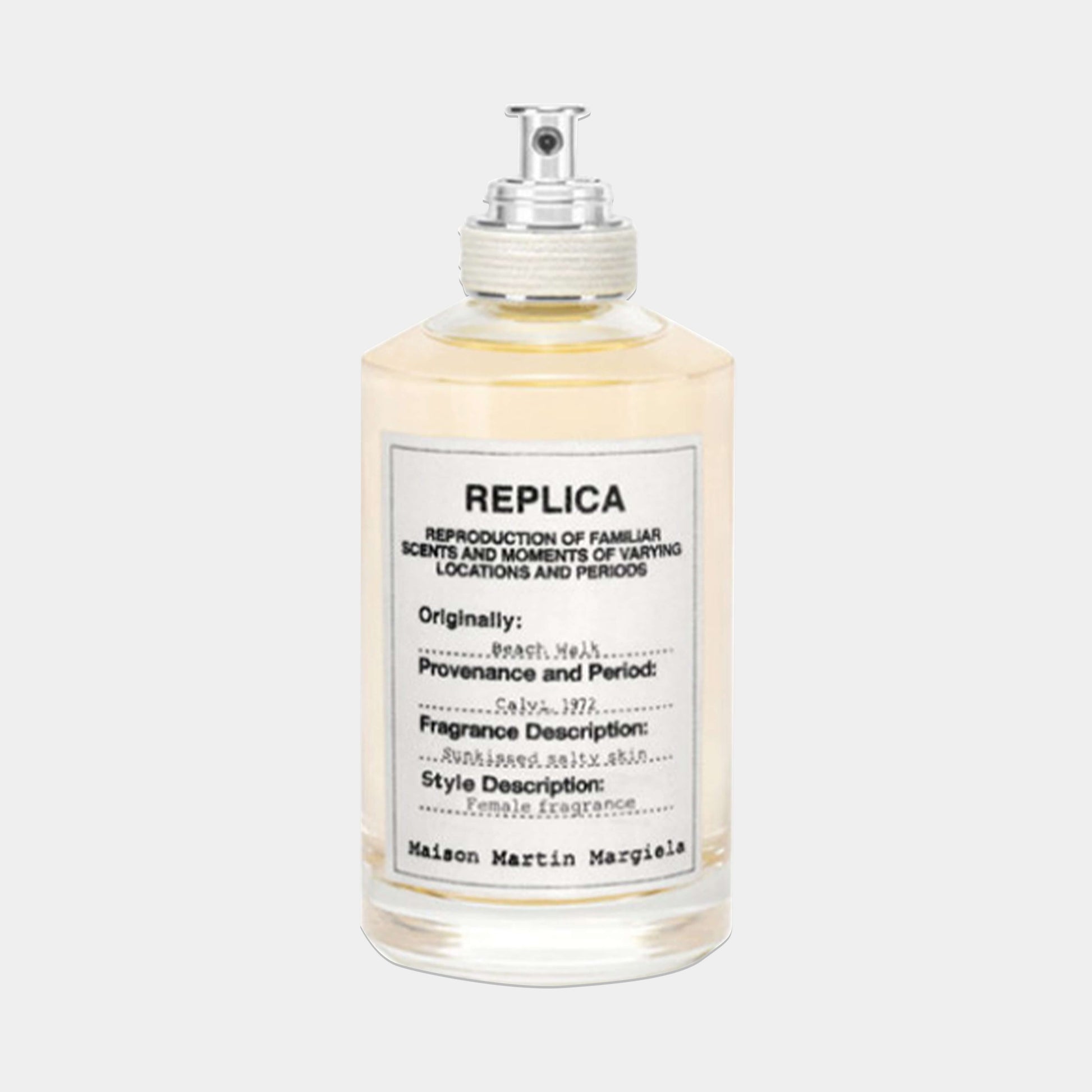 A bottle of Maison Martin Margiela Replica Beach Walk Sample perfume, featuring a clear glass design with a silver cap and a label detailing its fragrance description and origins. The liquid inside has a light yellow hue, evoking a sunny beach atmosphere.