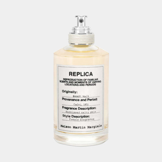 A bottle of Maison Martin Margiela Replica Beach Walk Sample perfume, featuring a clear glass design with a silver cap and a label detailing its fragrance description and origins. The liquid inside has a light yellow hue, evoking a sunny beach atmosphere.