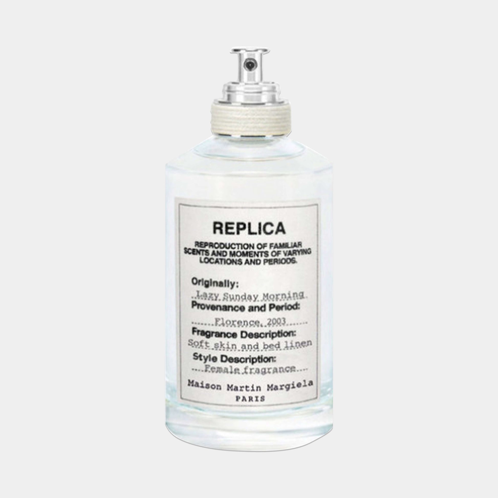 A clear glass bottle of Maison Martin Margiela Replica Lazy Sunday Morning Sample fragrance, featuring a minimalist label that describes its soft, floral scent inspired by fresh linen.