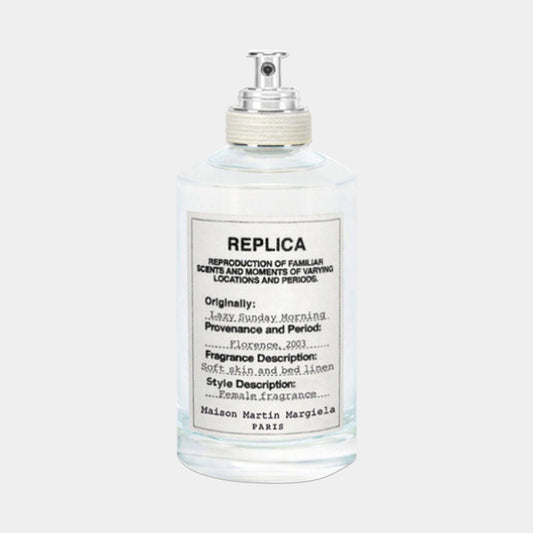 A clear glass bottle of Maison Martin Margiela Replica Lazy Sunday Morning Sample fragrance, featuring a minimalist label that describes its soft, floral scent inspired by fresh linen.