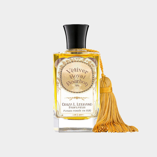 A glass bottle of Oriza L. Legrand Vetiver Royal Bourbon perfume, featuring a decorative yellow tassel and a vintage-inspired label. The liquid inside is a warm amber color, highlighting its luxurious appeal.