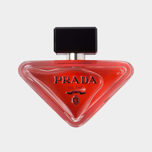 A stylish bottle of Prada Paradoxe Intense EDP, featuring a unique triangular shape and a vibrant red hue, topped with a black cap. The label displays “Prada Milano” prominently.