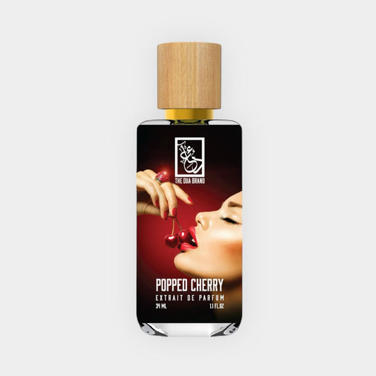 A bottle of Dua Popped Cherry Sample perfume, featuring a sleek design with a wooden cap and an artistic label depicting a woman holding cherries. The bottle contains 3 ml of fragrant extrait de parfum.